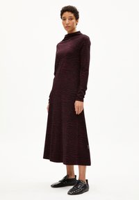 Knielanges Kleid in dunklem Burgunderrot mit Zebramuster, ausgestattet mit langen Ärmeln und hochgeschlossenem Hals. Getragen mit schwarzen, mit Nieten verzierten Schuhen.