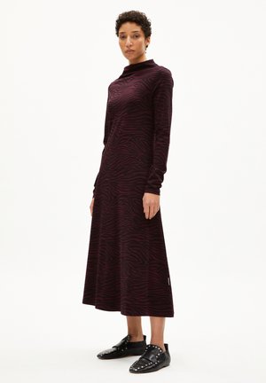 Knielanges Kleid in dunklem Burgunderrot mit Zebramuster, ausgestattet mit langen Ärmeln und hochgeschlossenem Hals. Getragen mit schwarzen, mit Nieten verzierten Schuhen.