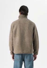 Tan fleecejakke med høj krave, der har en struktureret, blød overflade og en kontrasterende grøn indre krave, parret med blå denimjeans.
