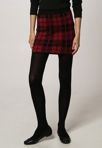 Femme portant une jupe écossaise rouge et noire, des collants noirs, des ballerines noires et un haut noir à manches longues, se tenant avec les mains détendues sur les côtés.