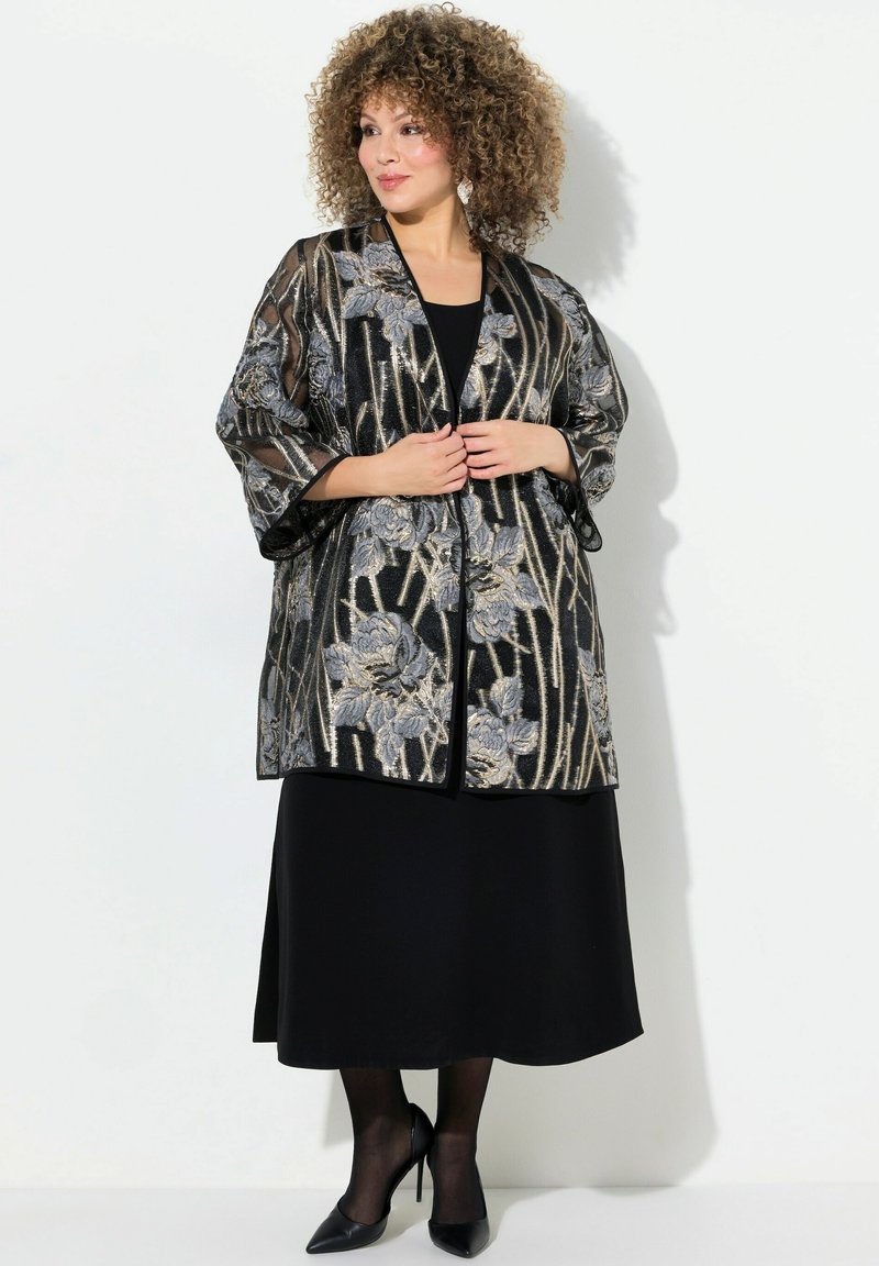 Schwarze Kimono-Jacke mit Blumenmuster aus transparenter Stoff, metallischen Akzenten, weiten Ärmeln und einem lässigen Schnitt, über einem schwarzen Kleid getragen.