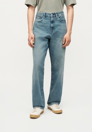 Widok z przodu osoby noszącej jasnoniebieskie jeansy o prostych nogawkach, beżowo-zieloną koszulkę i białe trampki z gumowymi podeszwami.