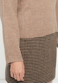 Beige gestrickter Pullover mit gerippter Textur, kombiniert mit einem Rock im Schwarz-Beige-Hahnentrittmuster. Eine Hand, die sichtbar auf dem Rock ruht.