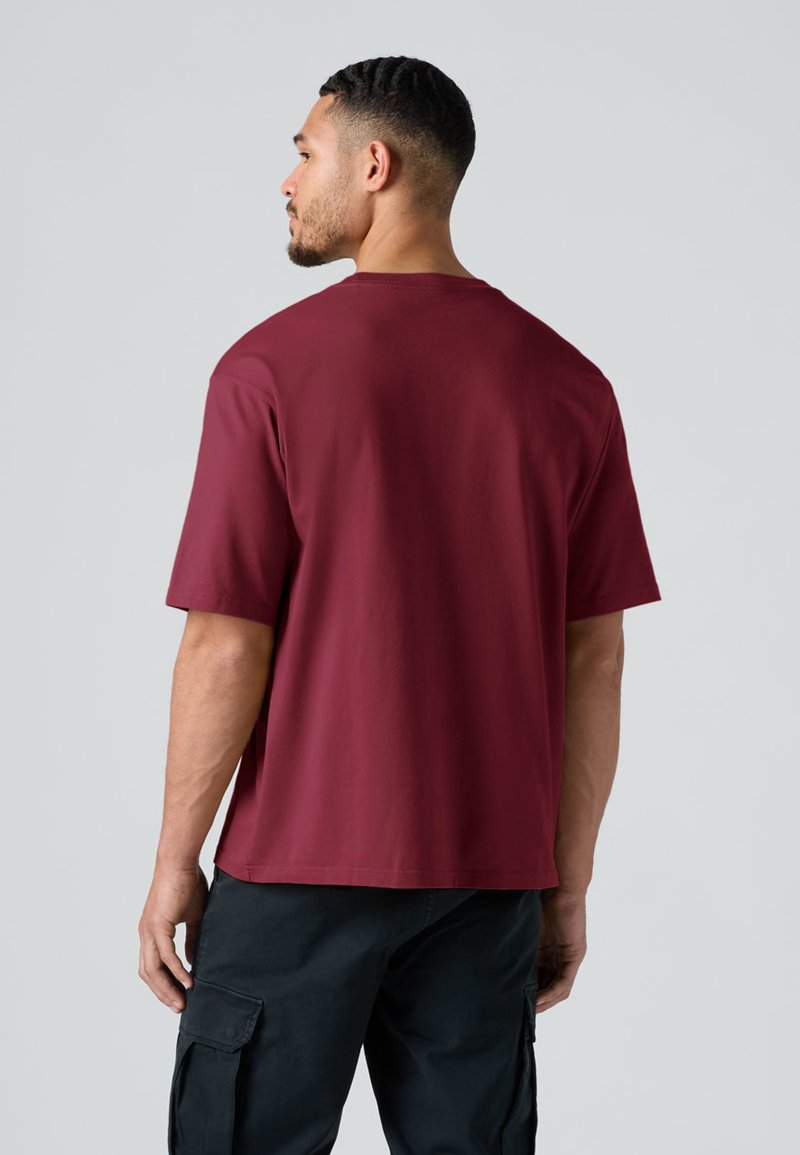 Uomo con capelli corti e barba che indossa una maglietta color ruggine semplice e pantaloni cargo neri, visto di spalle contro uno sfondo grigio chiaro.