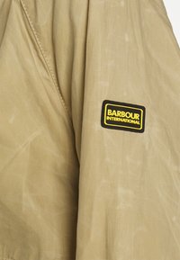 Khaki bunda vyrobená z měkké, texturované látky s jemným leskem. Obsahuje černou a žlutou nášivku "Barbour International" na rukávu.