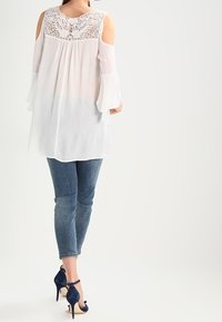 Blouse blanche à épaules dénudées avec des détails en dentelle, manches fluides, associée à un jean bleu ajusté et des escarpins navy.