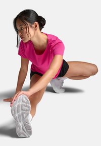 Conjunto deportivo que incluye una camiseta de ajuste ceñido y color rosa que absorbe la humedad y unos pantalones cortos negros, persona estirándose sobre un fondo blanco, usando zapatillas blancas.