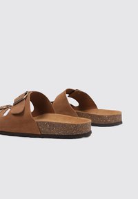 Sandalias deslizantes de ante marrón con detalles recortados y una plantilla de corcho. Suela de goma negra gruesa que proporciona agarre. Diseño simple, con parte trasera abierta para fácil uso.