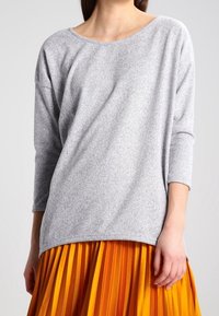 Grå, långärmad topp med vid halsringning och mjuk textur, ihop med en ljusorange plisserad kjol. Outfiten visar en kontrast i färg.