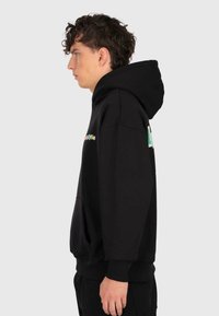 Jeune homme aux cheveux bouclés portant un sweat à capuche noir surdimensionné avec du texte coloré sur la poitrine, se tenant de profil contre un fond uni.