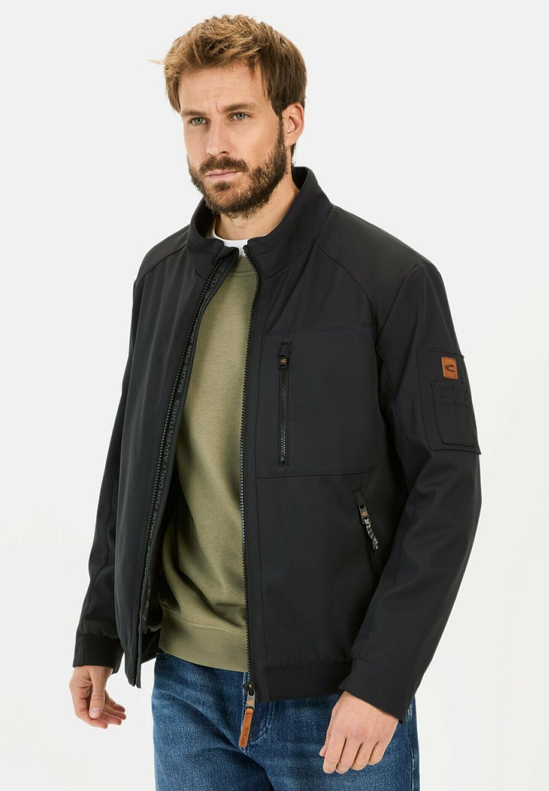 Sort jakke med høj hals, lynlåsfortil, sidelommer og ærmelap. Båret over en olivengrøn sweater og blå jeans.
