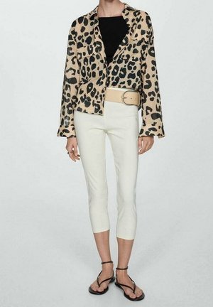 Personne portant une chemise boutonnée à imprimé léopard, un haut noir, un pantalon blanc court avec une ceinture beige et des sandales noires à lanières, debout sur un fond uni.