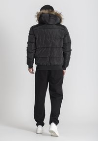 Giubbotto puffer nero con cappuccio foderato in pelliccia, dotato di sezioni trapuntate e polsini a coste. Indossato con pantaloni neri e sneakers bianche.