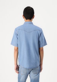 Šviesiai mėlynas džinsinis marškinėliai trumpomis rankovėmis, su smailiu apykakle, nugaros siūlės jungtimi ir subtiliais siuvinėjimo detalėmis.