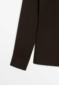 Chemise marron foncé à manches longues avec un poignet simple et des coutures propres sur fond blanc.