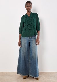 Blusa verde con davanti arricciato, maniche 3/4, abbinata a jeans in denim sbiadito a gamba larga. Modella che indossa stivaletti marroni alla caviglia. Sfondo semplice.