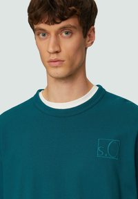 Teal crewneck sweatshirt met een gestructureerde logo patch op de borst, gemaakt van zachte stof; heeft een gelaagde witte halslijn.