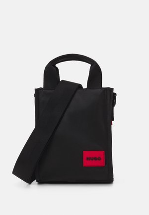 Schwarze Stofftasche mit kurzen Griffen und einem abnehmbaren Schultergurt, ausgestattet mit einem auffälligen roten HUGO-Logopatch auf der Vorderseite.