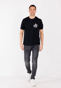 Man loopt vooruit en draagt een zwart T-shirt met een vogelafbeelding en tekst, donkergrijze jeans en witte sneakers, op een witte achtergrond.