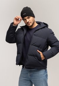 Granatowa kurtka puchowa z kapturem, z zapięciem na zamek i kieszeniami bocznymi. Połączona z czapką z prążkowanego materiału w kolorze czarnym i ciemnym swetrem.