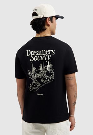 Mann trägt schwarzes T-Shirt mit dem Schriftzug "Dreamers Society" und einer illustrierten Dinner-Tafel mit Kerzen und Weinflaschen auf der Rückseite.