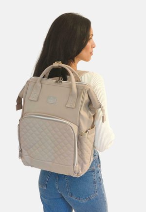 Mochila de quilt en beige con doble asa, un bolsillo frontal con cremallera y correas ajustables para los hombros. Tela suave con acentos plateados.