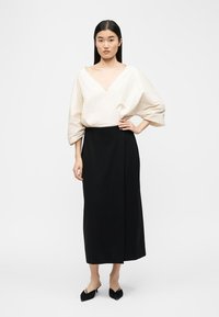 Γυναίκα με μακριά σκούρα μαλλιά που φοράει λευκό μπλουζάκι με λαιμόκοψη τύπου V και φουσκωτά μανίκια, μαύρη midi φούστα και μαύρες ψηλοτάκουνες μυτερές γόβες, στέκεται μπροστά σε λευκό φόντο.