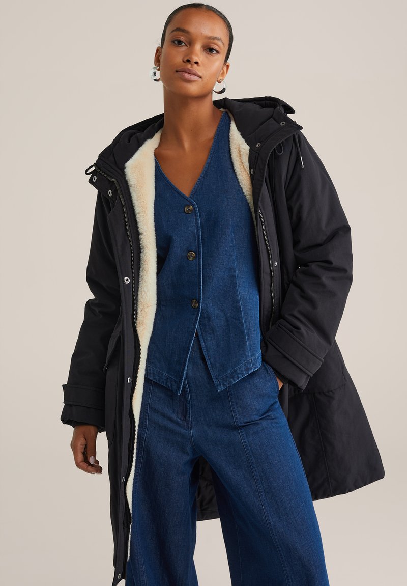 Zwart geïsoleerd parka met een crèmekleurige faux fur voering, ritssluiting en verstelbare capuchon, gedragen over een blauwe denim top en broek.