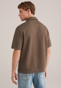Camisa polo marrón de manga corta con un patrón texturizado y un cuello, combinada con jeans de color azul claro. La camisa tiene un corte relajado y bordes acanalados.