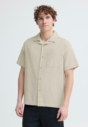 Mladý muž s kudrnatými vlasy, který má na sobě béžovou krátkou košili s knoflíky a náprsní kapsou, stojící před jednobarevným světlým pozadím.