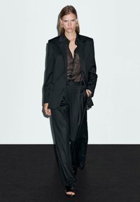 Black oversized blazer and tailored pants made of a smooth fabric, paired with a dark, sheer button-up shirt and black heeled sandals.