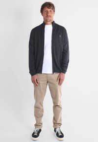 Veste zippée gris foncé sur un t-shirt blanc, chinos beiges et baskets noires avec semelles blanches. Le tissu semble doux et décontracté.