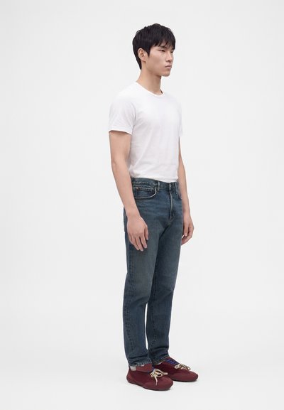Jeune homme debout, portant un t-shirt blanc uni, un jean bleu et des baskets bordeaux avec des lacets beiges, sur fond blanc.
