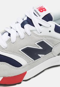 Sneaker grigio in suede e mesh con accenti blu navy, suola in gomma bianca e logo "N" evidente. Presenta lacci bianchi e dettaglio del tallone rosso.