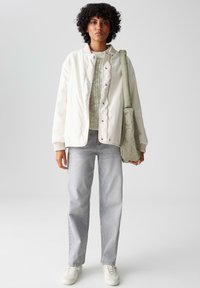 Witte geïsoleerde jas met eronder een gestructureerde groene trui, grijze spijkerbroek en een lichtgroene tas, gecombineerd met witte sneakers.