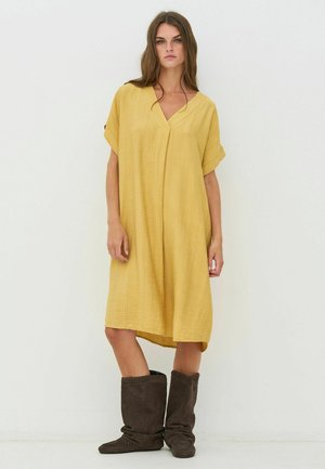 Vestito giallo, ampio, con maniche corte, scollo a V e tessuto strutturato. Abbinato a stivali marroni, morbidi, alti fino al ginocchio.