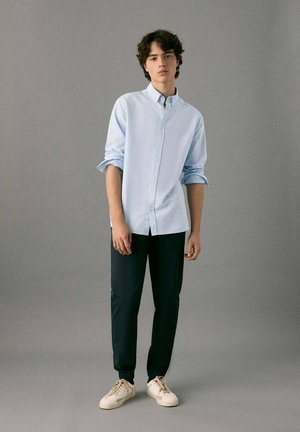 Chemise boutonnée bleu clair, pantalon noir et baskets crème. La chemise a un col et des manches retroussées. Design minimaliste sans motifs visibles.
