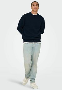 Maglione a girocollo blu navy con polsini e orlo a costine, abbinato a jeans chiari strappati e sneakers color crema.