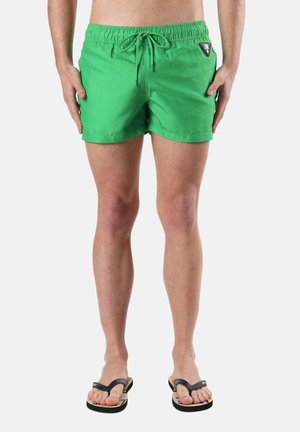 Homme portant un short de bain vert vif avec taille élastique et cordon, debout pieds nus en tongs noires sur un fond uni.
