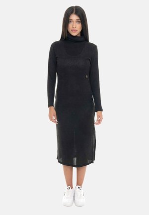 Donna in piedi, rivolta in avanti, che indossa un lungo vestito nero a collo alto con spacchi laterali e sneakers bianche su uno sfondo semplice.