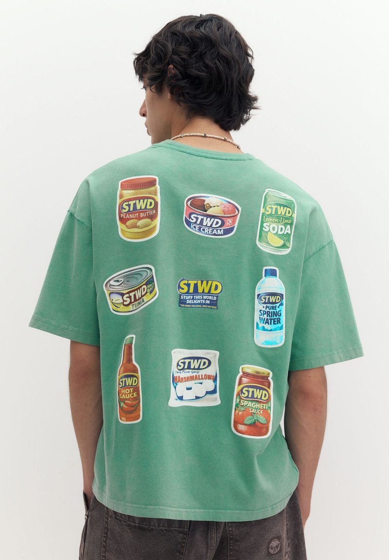 Jeune homme aux cheveux foncés porte une chemise verte affichant des illustrations colorées de produits alimentaires et de boissons dans le dos sur un fond uni.