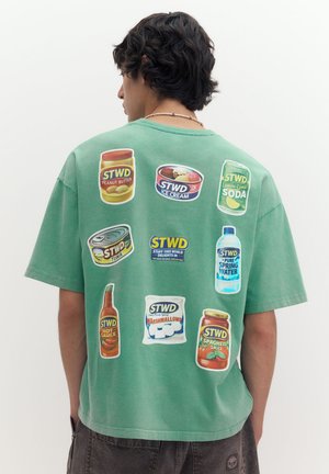 Jeune homme aux cheveux foncés porte une chemise verte affichant des illustrations colorées de produits alimentaires et de boissons dans le dos sur un fond uni.