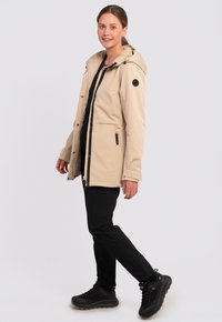 Beige, vandafvisende jakke med hætte, sort lynlås, sidelommer og sorte detaljer. Bæres over en sort top og kombineres med sorte bukser.