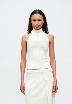 Sötét, göndör hajjal rendelkező nő ujjatlan, fehér, bordázott garbós felsőt és hozzá illő, magas derékú szoknyát visel, egy sima, világos háttér előtt.