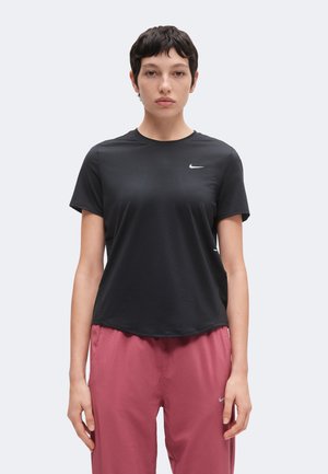 Άτομο με κοντά σκούρα μαλλιά φοράει μαύρο t-shirt Nike και ροζ παντελόνι, στέκεται κοιτώντας μπροστά σε ένα απλό ανοιχτόχρωμο φόντο.