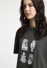 Camiseta de algodón verde oscuro con cuatro estampados gráficos en blanco de plantas estilizadas y diseños en la parte delantera. Cuello de tripa acanalado.
