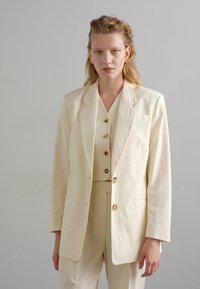 Licht beige textuur pak bestaande uit een blazer met notch revers, een aansluitende vest met knopen en bijpassende broek.