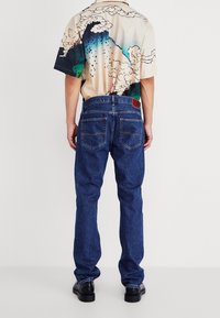 Persona con vaqueros azules y una camisa de manga corta con un patrón colorido de olas y nubes, de pie de espaldas sobre un fondo liso.