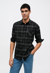 Camisa xadrez preta e cinza com mangas compridas, colarinho com botões e bolso no peito, combinada com jeans azuis. Textura do tecido lisa.