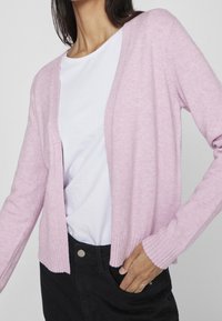 Helles rosa Cardigan aus weichem Stoff, mit V-Ausschnitt, offener Vorderseite und gerippten Ärmelbündchen. Getragen über einem weißen Hemd und schwarzen Hosen.